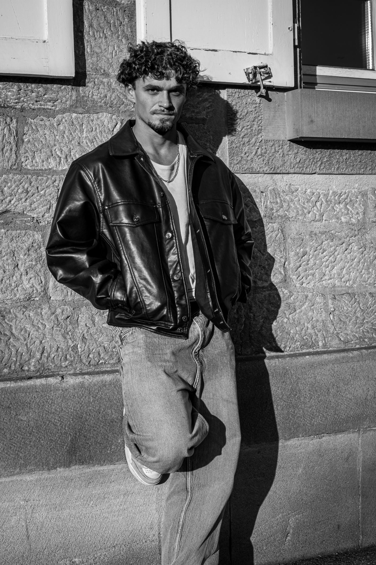 Black and white portrait of a man in a leather bomber jacket leaning against an urban wall