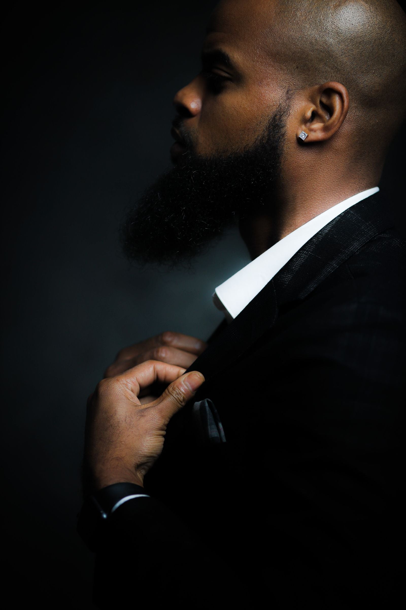 Close-up side profile of a man adjusting a tailored black suit blazer against a dark background